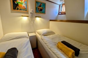 Zweibettkabine mit zwei Einzelbetten, weißer Bettwäsche, gelben und schwarzen Handtüchern, Wandlampen, bunten Bildern und kleinem Fenster auf Floor van Aemstel.