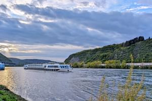 Weißes Flusskreuzfahrtschiff SE Manon auf dem Rhein bei Andernach, mit grünen bewaldeten Hügeln auf beiden Seiten unter bewölktem Abendhimmel.