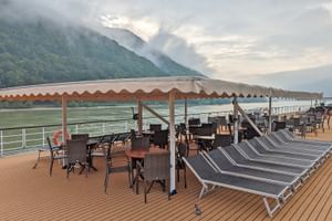 Sonnendeck der MS Prinzessin Katharina mit Liegestühlen, Tischen und Sitzgelegenheiten unter Markisen. Fluss und bewaldete Berge im Hintergrund.