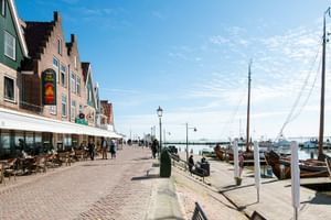 Uferpromenade in Volendam mit traditionellen holländischen Backsteinhäusern, Café-Terrassen und Holzbooten im Hafen unter blauem Himmel.