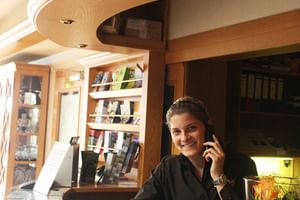 Lächelnde Rezeptionistin am Telefon an der Holzrezeption des Hotel Central Zeven mit geschwungener Decke und Produktregalen im Hintergrund.