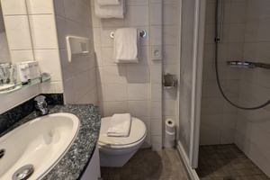 Kompaktes Badezimmer auf der MS Olympia mit weißem Waschbecken, Granitplatte, Toilette, ebenerdiger Dusche mit Glastür und weißen Handtüchern.