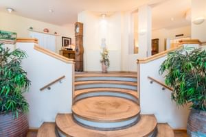 Heller Hoteleingang mit geschwungener Treppe, Holzgeländern, großen Topfpflanzen und Standuhr. Warmes Licht schafft einladende Atmosphäre.