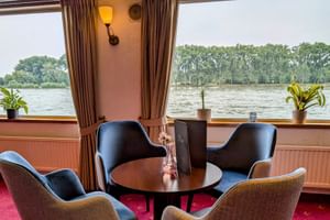 Gemütlicher Salon an Bord der MS Olympia mit vier Stühlen um einen Tisch, großen Fenstern mit Fluss- und Baumblick, braunen Vorhängen und rotem Teppich.