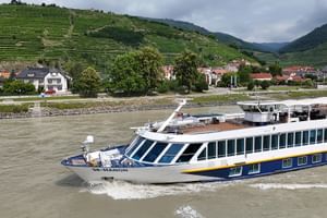 Flusskreuzfahrtschiff SE Manon mit blau-gelbem Rumpf fährt auf der Donau. Terrassierte Weinberge und ein Dorf auf Hügeln im Hintergrund.