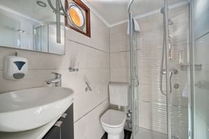 Kompaktes Magellan Schiff Badezimmer mit weißem Waschbecken, Toilette, Glasduschkabine, Spiegel und Bullauge mit Holzrahmen.