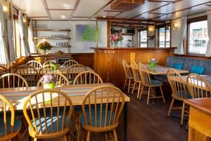 Interior of MS Flora's salon restaurant with wooden tables, Windsor chairs, blue banquette seating, and a service counter with nautical decor.
