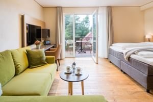Modernes Hotelzimmer mit grünem Sofa, Doppelbett, TV und Balkontür mit Blick auf Sitzbereich und Garten.