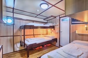 San Snova Schiffskabine mit drei Betten: Etagenbett links, Einzelbett rechts. Weiße Wände, Holzböden, Bullaugen und Schließfächer.