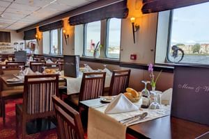 Innenraum des MS Normandie Schiffsrestaurants mit gedeckten Tischen, Holzstühlen und großen Fenstern mit Flussblick. Roter Teppich und warme Beleuchtung.