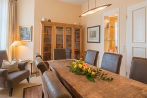 Elegantes Esszimmer mit Holztisch, grauen Stühlen, frischem Blumenarrangement, Holzbücherschrank, Sessel und beleuchtetem Vitrinenschrank.