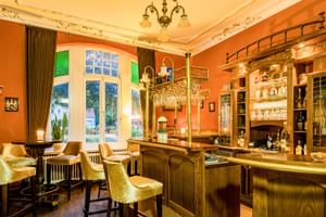 Elegante Hotelbar mit orangefarbenen Wänden, goldenem Holztresen, Messingzapfhähnen, Samtstühlen und verzierter Decke. Große Fenster mit Vorhängen.