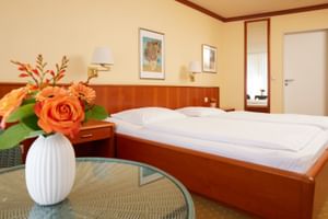 Helles Hotelzimmer mit Doppelbett, Holzmöbeln, weißer Vase mit orangen Rosen auf Glastisch und cremefarbenen Wänden.