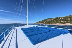 Weißes Sonnendeck der San Snova mit blauer Sitzfläche, weißen Geländern, Mast und Blick auf blaues Meer und grüne Küste.