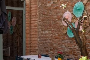 Außenterrasse mit weißem Tisch, bunten Stühlen und dekorativen Papierfächern an einem Baum vor einer Backsteinmauer in Toulouse.