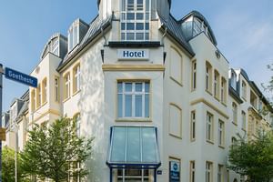 Weißes Eckgebäude mit markanter Turmspitze und blauem 'Hotel'-Schild. Hotel Geheimer Rat mit gelben Fensterrahmen und modernem Glaseingang.