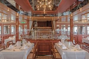 Elegantes Restaurant an Bord der MS Switzerland mit weiß gedeckten Tischen, Holzstühlen und einem zentralen Klavier. Warme Beleuchtung und Fenster.