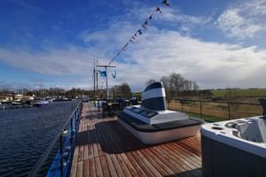 Wooden sun deck of MS MAGNIFIQUE IV with blue railings, funnel, and mast with colorful flags. Harbor and countryside visible in background.