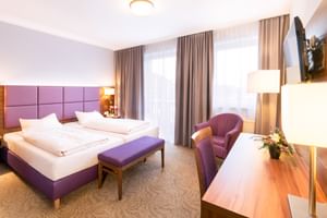Modernes Hotel Seeblick Doppelzimmer mit lila gepolstertem Kopfteil, weißer Bettwäsche, lila Sessel, Holzschreibtisch und grauen Vorhängen.