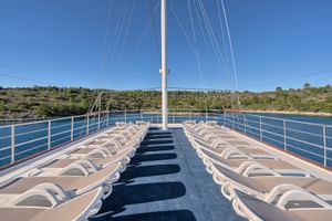 Sonnendeck der MY Magellan mit Reihen weißer Liegestühle auf blauem Teppich, weißen Geländern und Mast. Kroatische Küste mit blauem Wasser sichtbar.