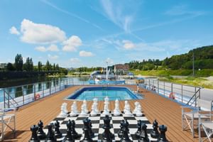Sonnendeck der SE Manon mit Swimmingpool, übergroßem Schachspiel auf Schachbrett, Holzdeck und Flussblick unter blauem Himmel.