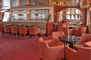 Elegante Salon Bar auf der MS Switzerland mit korallfarbenen Sesseln, runden Tischen und langer Tafel. Mit rotem Teppich und warmer Beleuchtung.