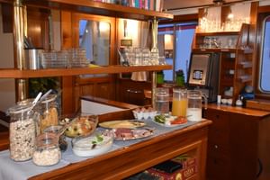 Breakfast buffet on MS Flora with cereals, cold cuts, cheese, juice and coffee machine in elegant wooden ship interior with glasses and plates.