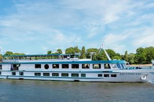 Weißes Flusskreuzfahrtschiff MS Switzerland mit blauem Zierstreifen auf ruhigem Wasser. Bäume und blauer Himmel im Hintergrund.