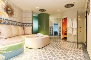 Moderner Wellness-Bereich mit geschwungener Sitzbank, grüner zylindrischer Dusche, geometrischen Bodenfliesen und hängenden Bademänteln.