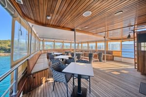 Geräumiges Außenlounge-Deck auf dem Schiff Magellan mit Holzdecke, Panoramafenstern, Esstischen, Stühlen und Meerblick.