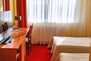 Helles Hotelzimmer mit zwei Einzelbetten, Holzschreibtisch, Stuhl, TV, rotem Teppich und weißen Gardinen mit orangen Vorhängen im Hotel Ludmila in Mělník.