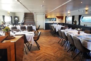 Elegant restaurant interior on MS Magnifique IV with white-clothed tables, dark chairs, parquet flooring, and panoramic windows.