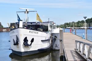 Weißes Flusskreuzfahrtschiff MS Fluvius mit schwarzen Ankern an einem Holzsteg festgemacht. Marina mit Segelbooten und Bäumen im Hintergrund.
