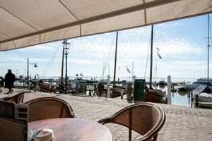 Überdachte Terrasse mit Holztischen und Korbstühlen mit Blick auf den Hafen von Volendam mit traditionellen Booten und IJsselmeer.