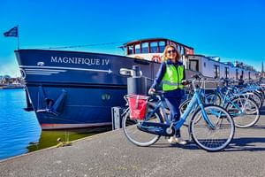 Woman in high-vis vest with bicycle standing beside MS Magnifique IV cruise ship moored at harbor under clear blue sky.