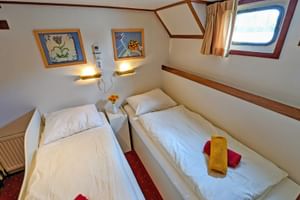 Zweibettkabine mit zwei Einzelbetten, weißer Bettwäsche, Bullauge, Wandlampen und gerahmten Bildern auf Floor van Aemstel Schiff.