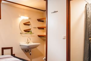 Kompaktes Badezimmer auf Segelschiff Antigua mit weißem Waschbecken, rundem Spiegel, Holzregalen und Duschbereich mit grauem Vorhang.