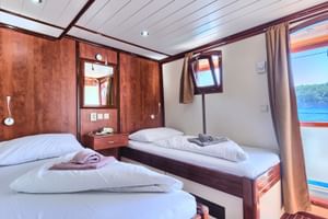 Twin cabin on MY Andela Lora with two single beds, white linens, wood paneling, and large window showing coastal view.
