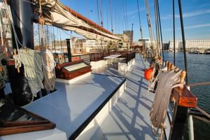 Weißes Deck des Segelschiffs Simmer fan Fryslân mit Holzbänken, Takelage, Masten und Hafengebäuden im Hintergrund unter blauem Himmel.