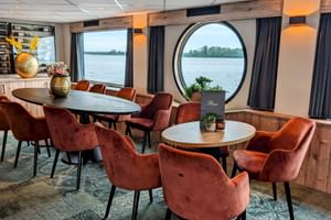 Moderne Lounge auf der MS Princess mit rostfarbenen Samtstühlen, ovalem Esstisch und großem rundem Fenster mit Flussblick.