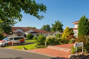 Weiße Hotelgebäude mit roten Ziegeldächern in Auefeld, umgeben von grünem Rasen und buntem Garten. Hotel-Shuttle vor dem Gebäude.