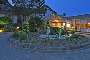 Landhotel Schnuck in der Dämmerung mit beleuchtetem Eingang und moderner Glasfassade. Runder Garten mit Skulptur und Zierpflanzen im Vordergrund.