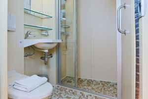 Kompaktes Badezimmer auf der SE Manon mit Waschbecken, Spiegelschrank, Glasregalen, begehbarer Dusche mit Mosaikfliesen und weißen Handtüchern.