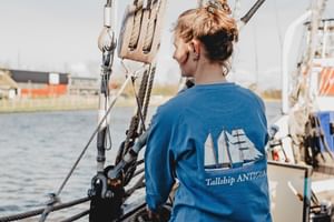 Person in blauem Sweatshirt mit Tallship Antigua Logo steht an Deck, umgeben von Takelage und Seilen, mit Hafengebäuden im Hintergrund.