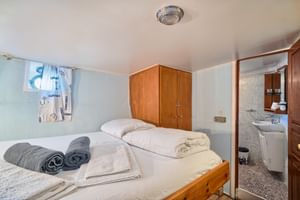 Kompakte Doppelkabine auf der Panagiota mit zwei Einzelbetten, weißer Bettwäsche, grauen Handtüchern, Holzschrank und eigenem Bad.