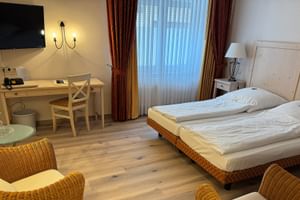Helles Hotelzimmer im Hotel Central Zeven mit Doppelbett, Holzschreibtisch, Korbstühlen, Flachbildfernseher und roten Vorhängen.