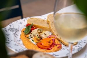 Dekorativer Teller mit Hummus, geschnittenem Gemüse, Kräutern und Brot, serviert mit einem Glas Weißwein auf einem Tisch im Freien.