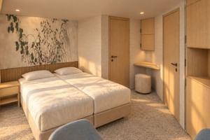 Modern suite on MS Veloria with twin beds, light wood furniture, botanical wall art, and beige carpet. Built-in wardrobes and vanity area visible.