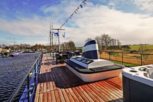 Holz-Sonnendeck der MS Magnifique Vier mit Whirlpool und Liegestühlen, Blick auf Marina mit Booten und grünen Feldern unter blauem Himmel.