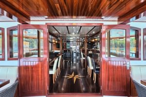 Interior view of MY Andela Lora ship salon with polished wooden floors, ceiling beams, dining tables, white seating, and large windows overlooking water.